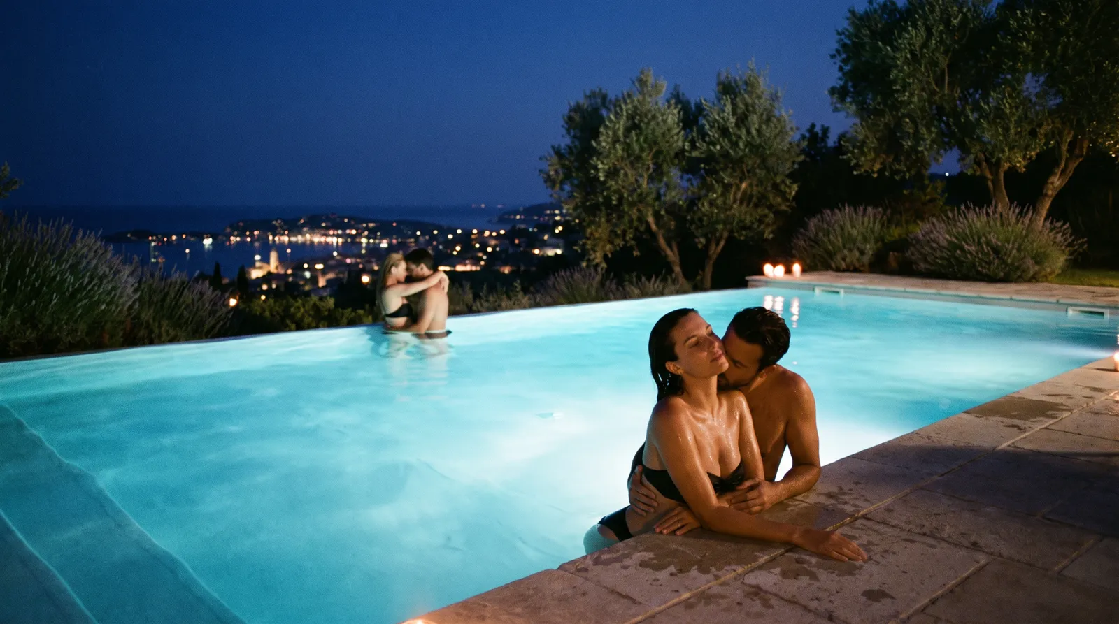 Couple enlacé au bord d'une piscine à débordement éclairée la nuit, au-dessus des lumières de la Côte d'Azur, un autre couple plus loin dans l'eau
