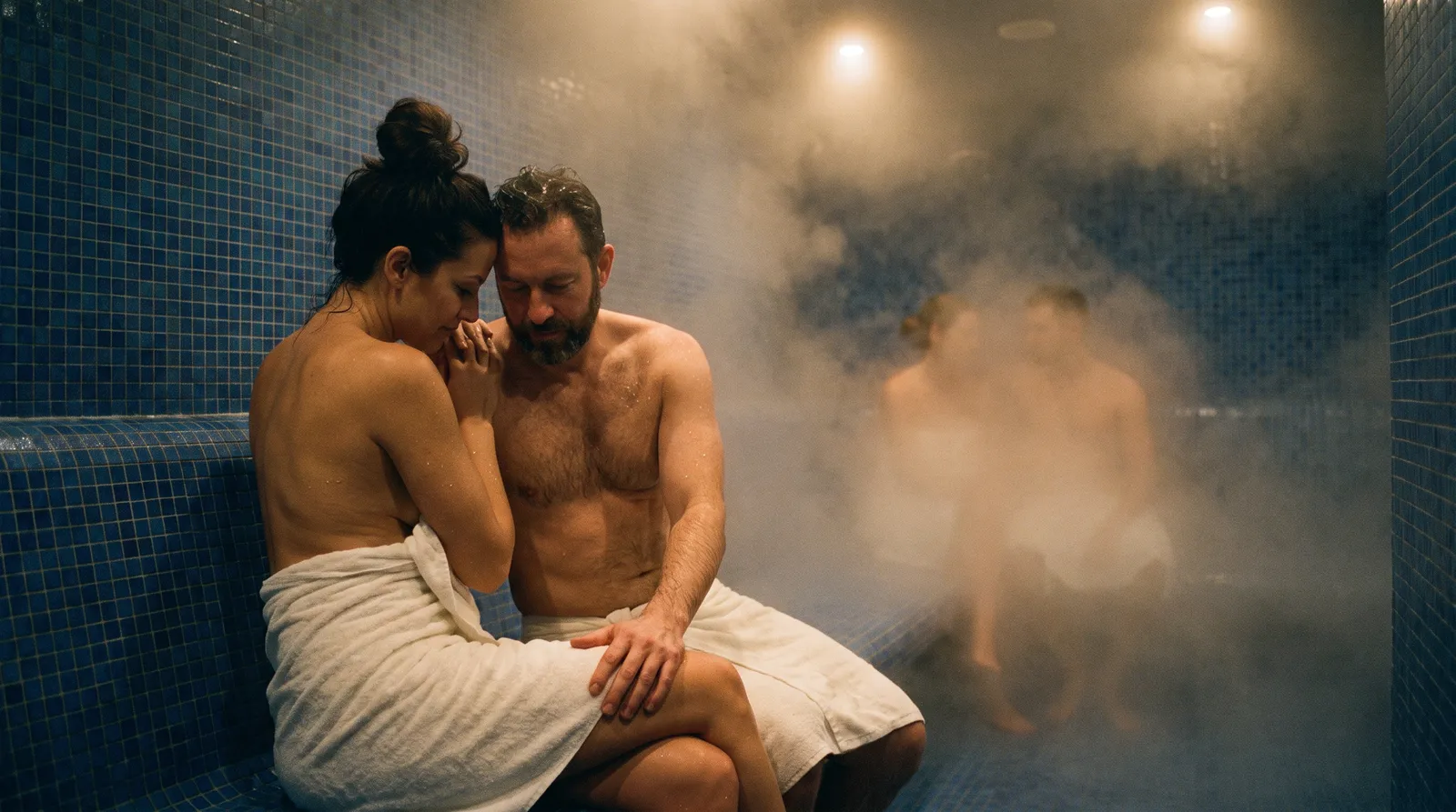 Couple en serviettes blanches serré l'un contre l'autre dans un hammam en mosaïques bleues, un autre couple dans la vapeur