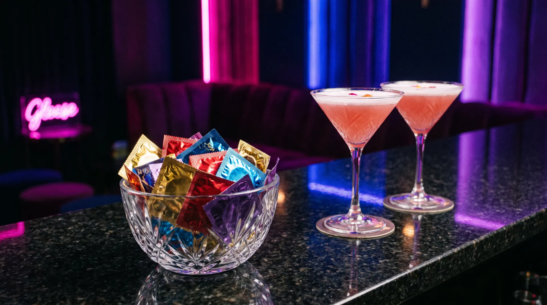 Coupelle de préservatifs posée sur le bar d’un club libertin, à côté de deux cocktails roses