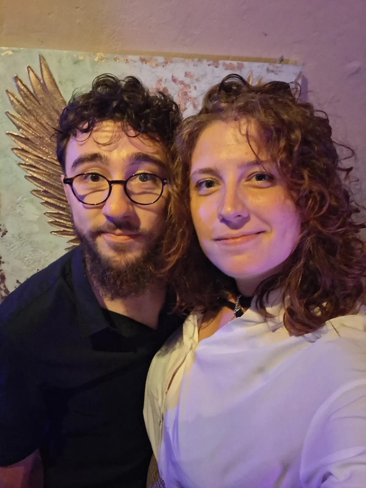 Selfie d’Antodeline : lui barbu à lunettes rondes, elle aux cheveux bouclés, devant une fresque aux ailes dorées