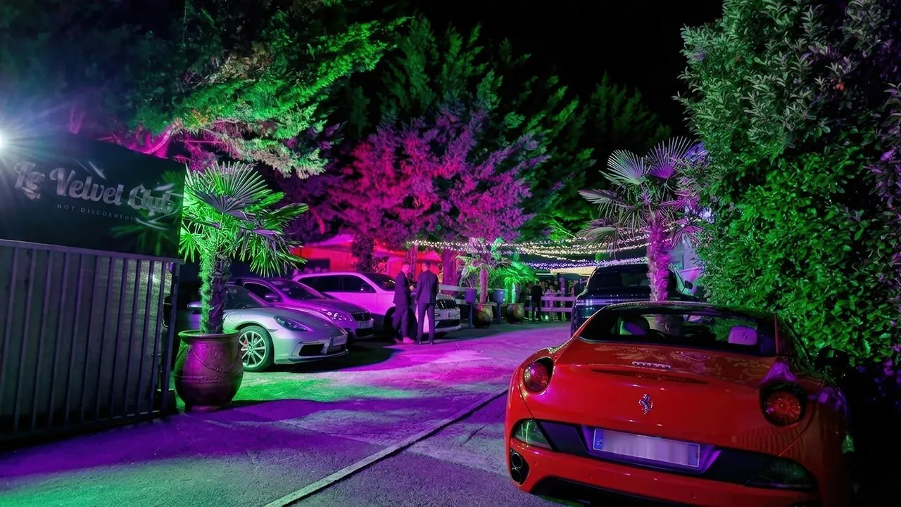 Le parking du Velvet Club la nuit, bordé de palmiers et d'arbres éclairés en vert et violet, devant l'entrée du club